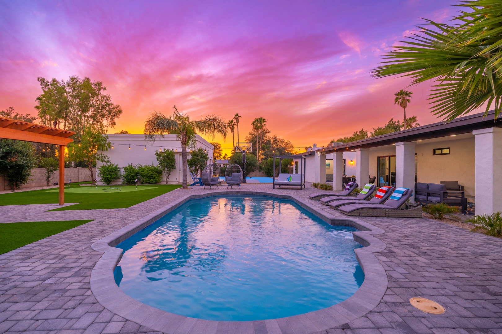 This stunning property features a sparkling pool, landscaped lawn, covered patio, and modern architecture—all framed by vibrant desert palms and breathtaking sunset skies painted in pink, purple, and gold.