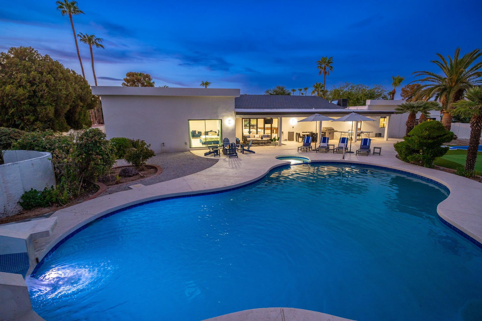 Modern desert retreat featuring a sparkling pool, covered patio seating, and lush palm trees under twilight skies—your perfect oasis for relaxation.