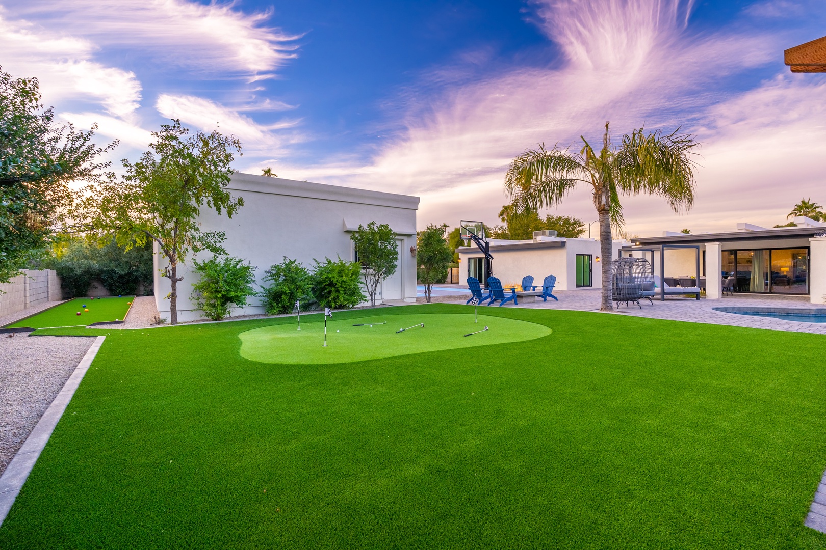 Modern property featuring a private putting green, basketball court, and pool area surrounded by palm trees and lush landscaping under vibrant desert skies.