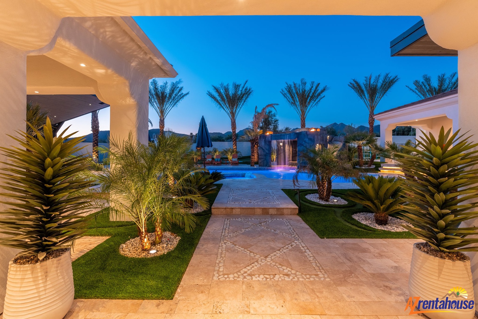 Stunning resort-style pool area framed by elegant archways, lush palm trees, and illuminated landscaping during twilight hours.
