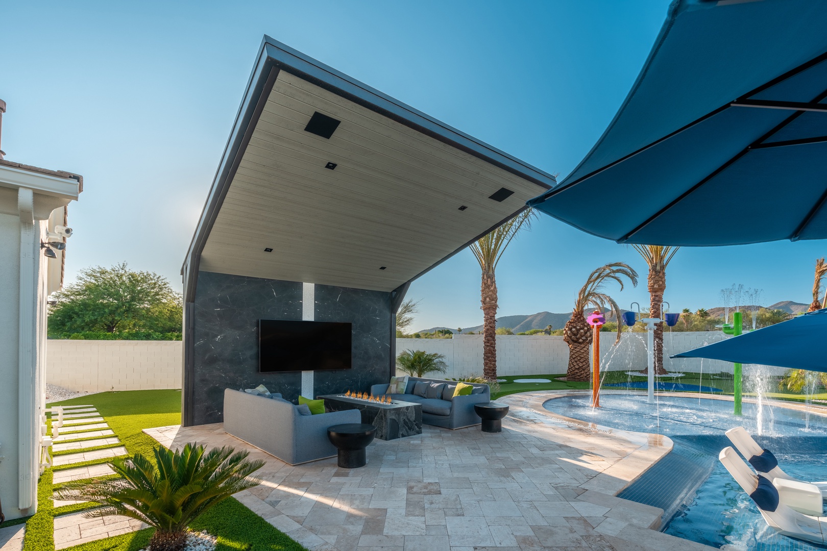 The property features a contemporary outdoor pavilion with an integrated fireplace, overlooking a resort-style pool equipped with water features and palm trees.