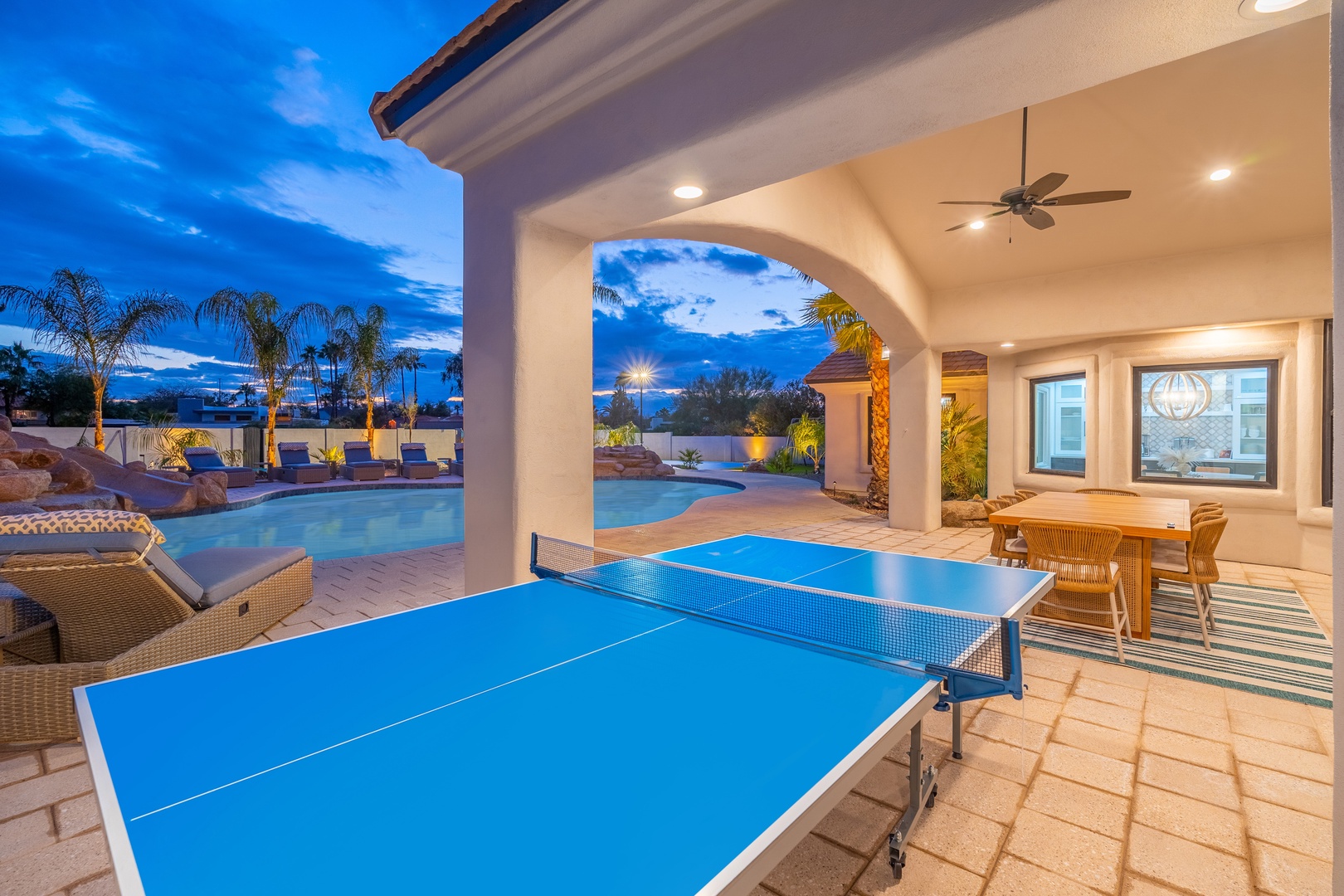 A covered patio area with table tennis setup overlooking the sparkling pool. Warm lighting and outdoor dining create the perfect space for evening entertainment under the stars.