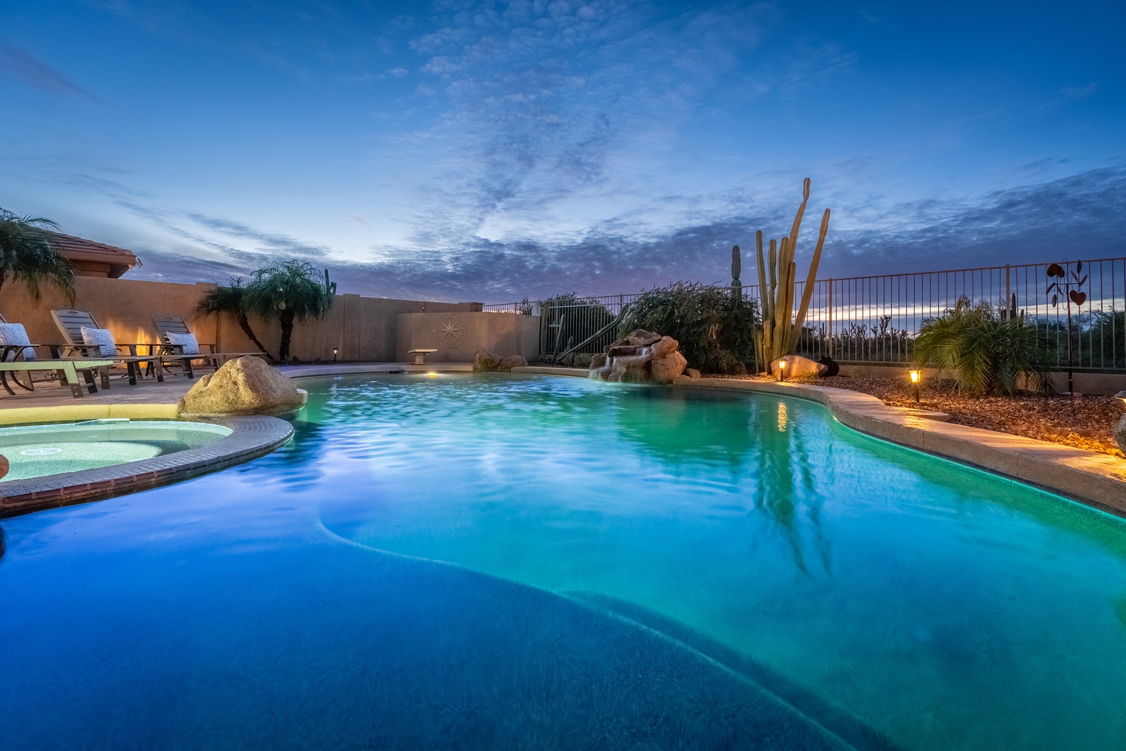 Picture yourself floating in this stunning desert oasis at twilight, where glowing pool lights dance beneath a painted sky. Natural rock features and native cacti frame your private swimming paradise.