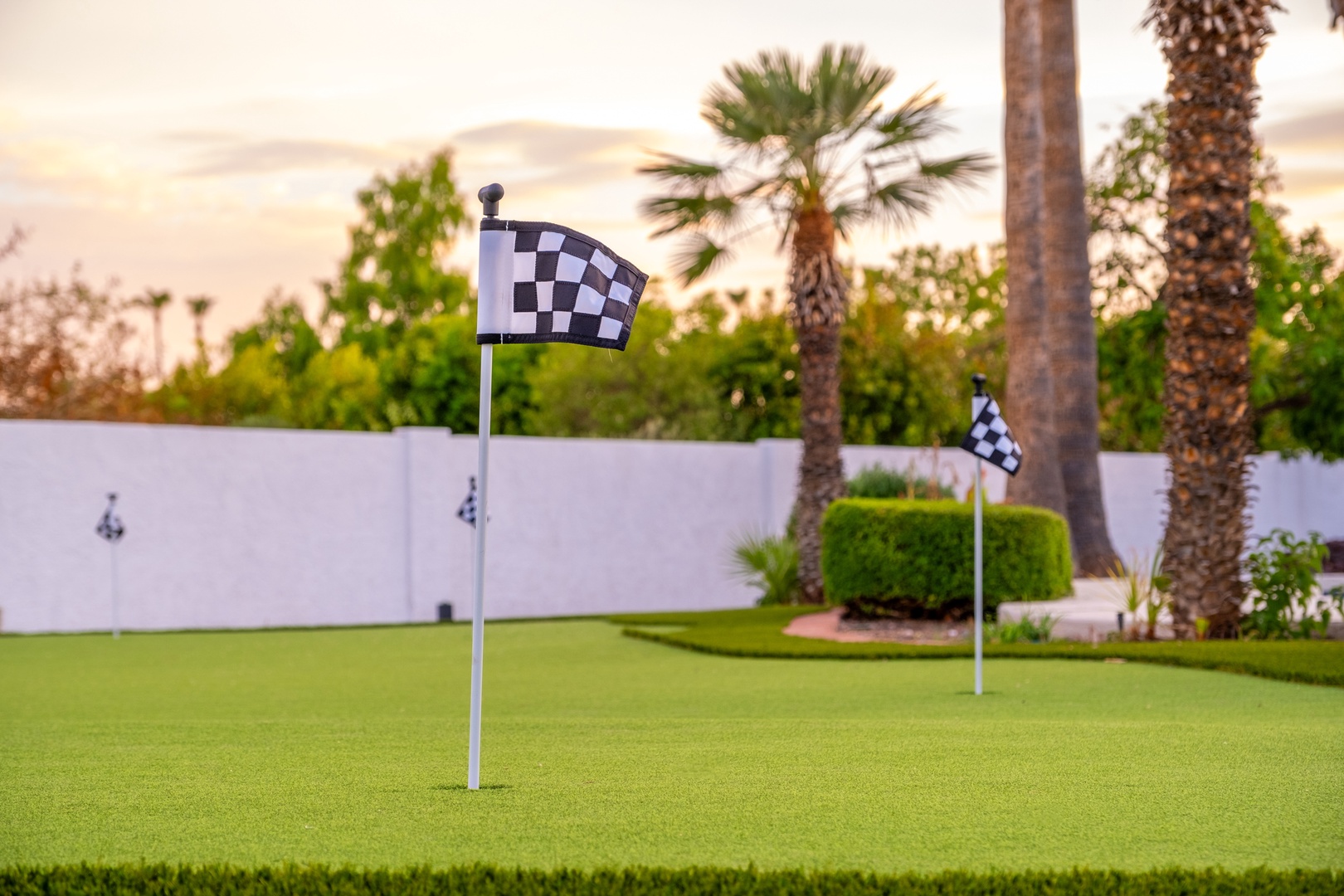 Private putting green features checkered flags amid manicured landscaping and palm trees, perfect for perfecting your short game in a tranquil backyard setting.