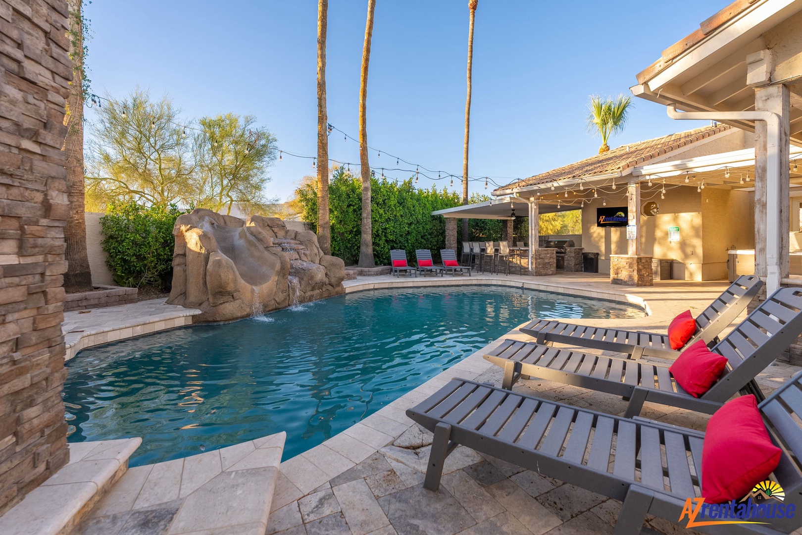 A private oasis featuring a sparkling pool framed by towering palms, stone waterfall, shaded patio, and multiple loungers for ultimate relaxation under desert skies.