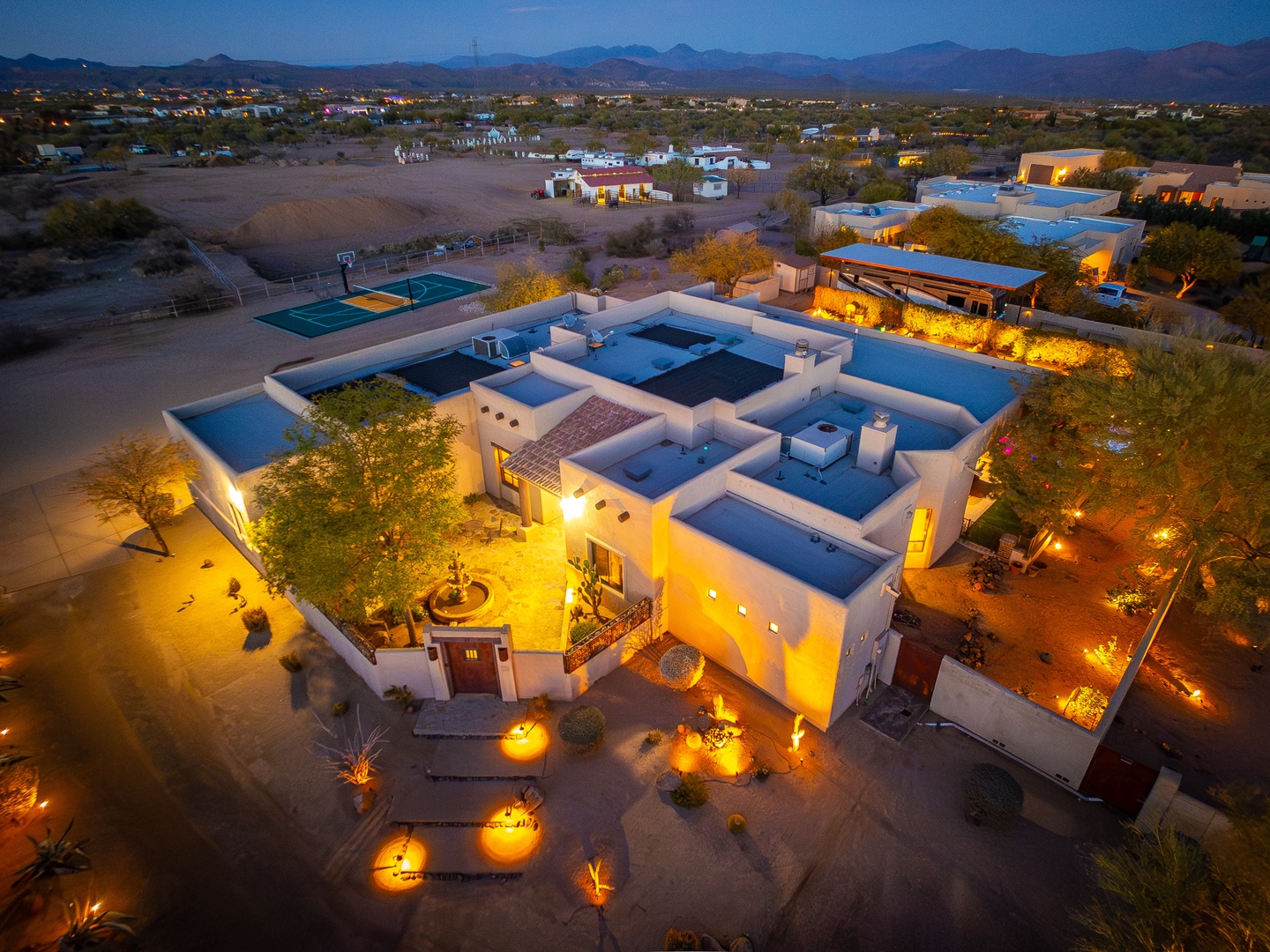 Stunning desert estate nestled in Arizona's Sonoran landscape, featuring Southwestern architecture, private tennis court, and dramatic mountain views. Property illuminated at twilight showcases extensive outdoor living spaces amid natural desert setting.