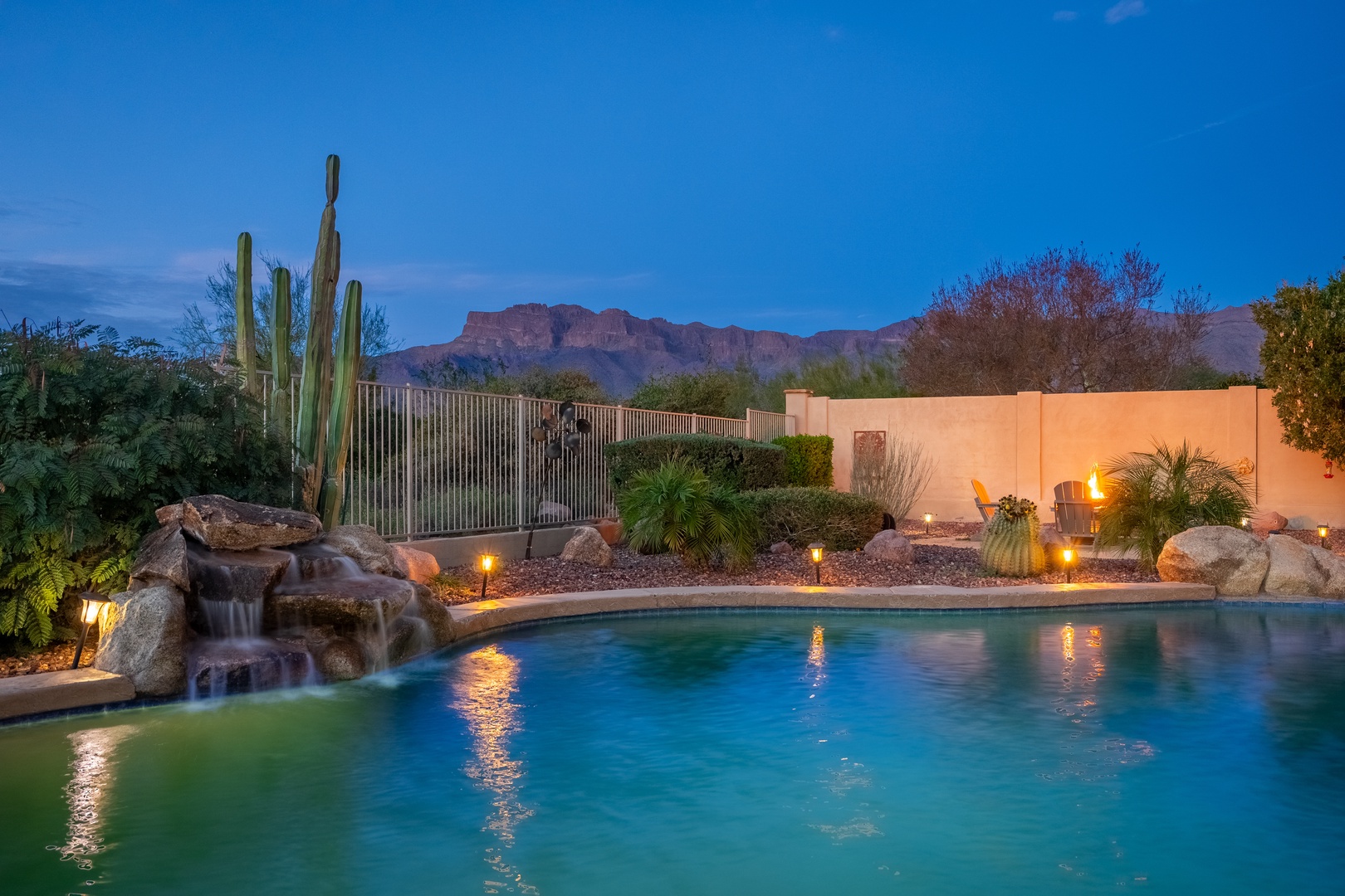 A stunning desert oasis at twilight, featuring a resort-style pool with rock waterfall, desert landscaping including saguaro cacti, and dramatic mountain backdrop under evening sky.