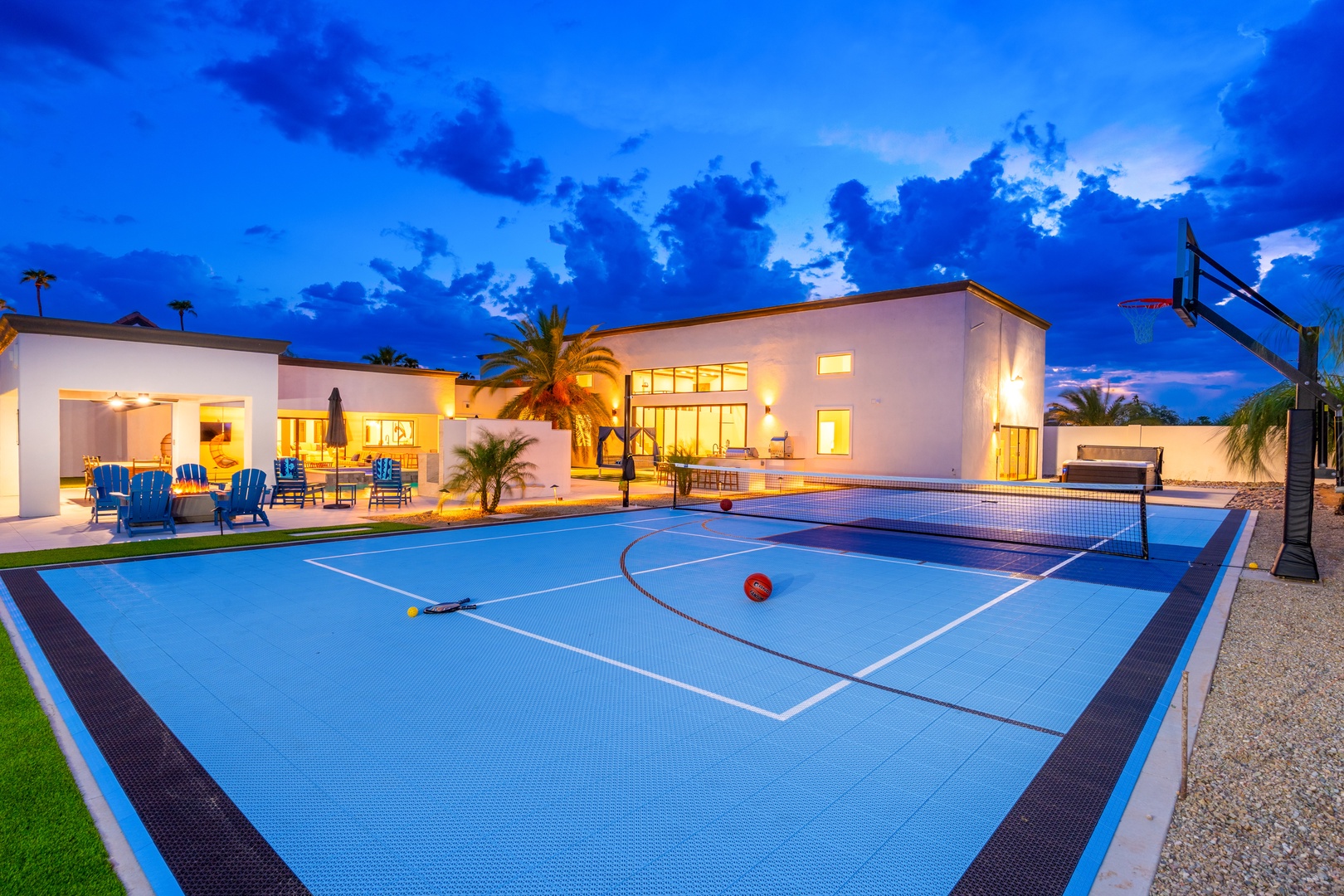 Modern desert retreat with a full-size tennis court, basketball hoop, and glowing outdoor spaces set against a dramatic evening sky.