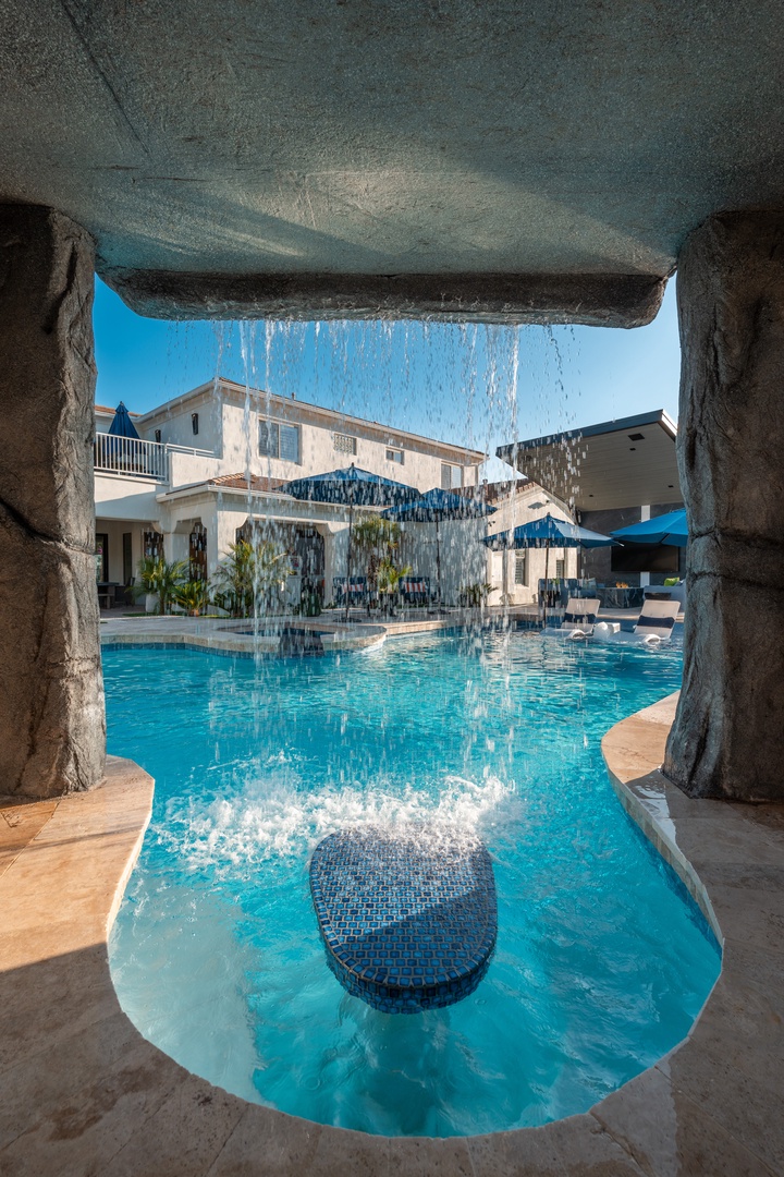 Stunning resort-style pool features a dramatic waterfall cascading through a rock grotto, creating your own private oasis under bright blue skies.