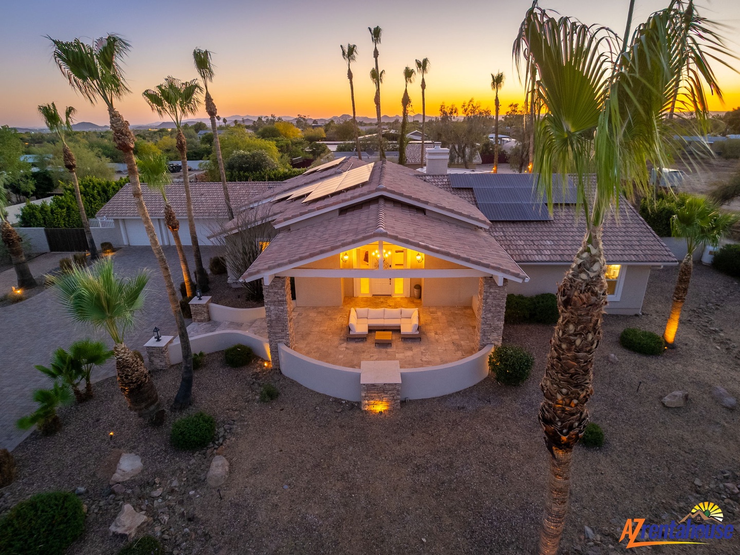 An elegant desert retreat with a covered patio and seating areas, surrounded by palm trees, captured at sunset with soft evening light illuminating the architecture and neighborhood beyond.