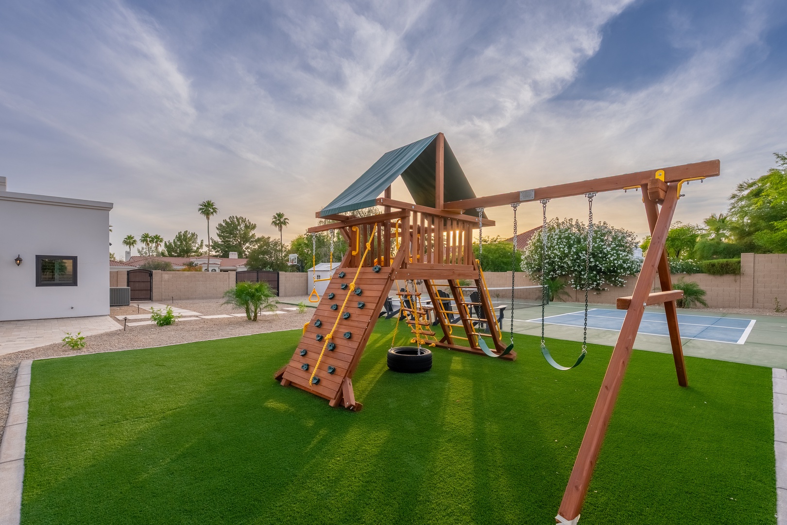 A spacious backyard features a wooden playground structure with climbing wall, swings, and slide on lush artificial turf, plus a sports court and scenic desert views.