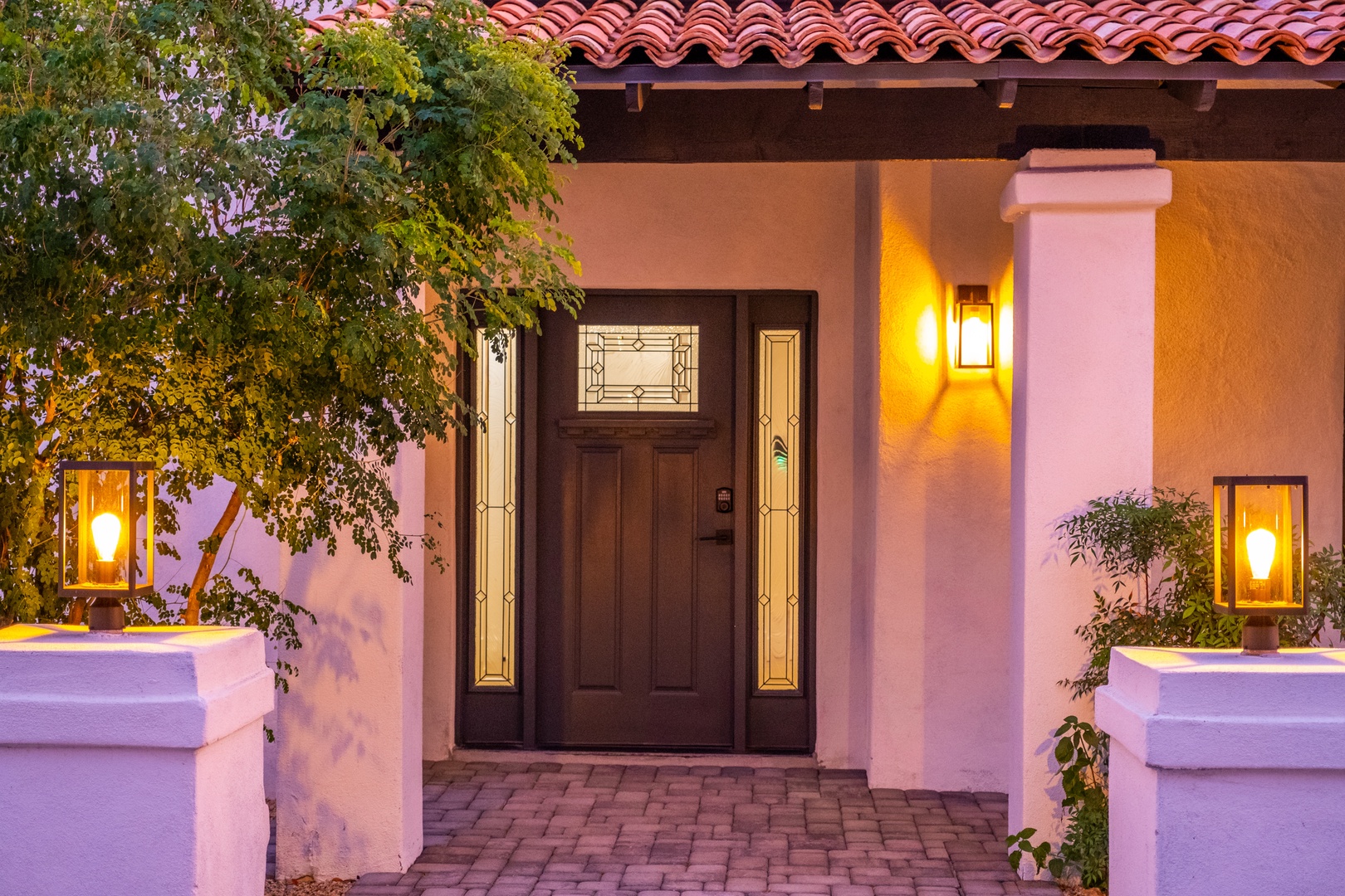 Welcome home to your Spanish-style retreat, where warm evening lighting illuminates the entrance through elegant pillars and red tile roof. The private paver walkway sets the scene for relaxation.