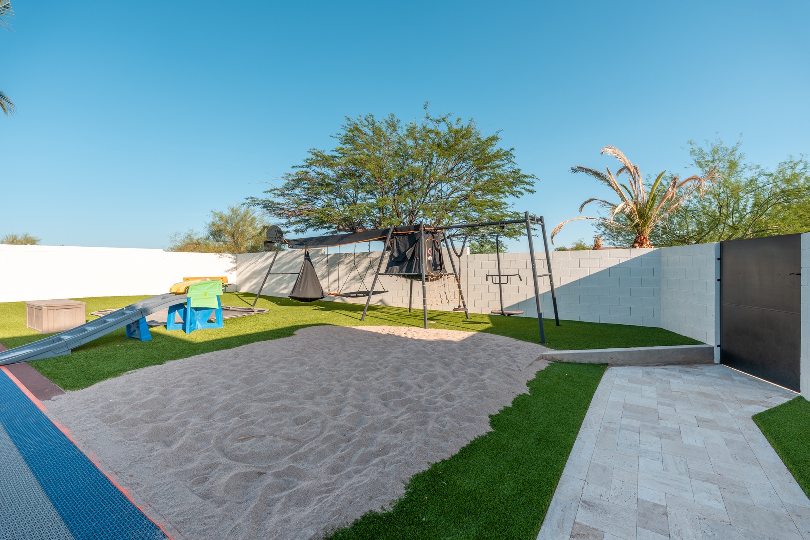 A private backyard designed for family fun features a swing set, sandbox, and soft turf, surrounded by lush green landscaping and clear blue skies.