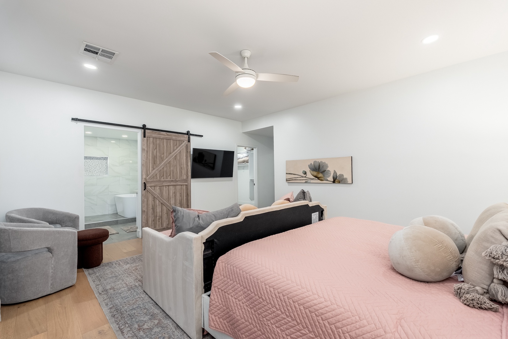 Unwind in your serene retreat with plush bedding, cozy seating, and a spa-like ensuite visible beyond the stylish barn door, all bathed in natural light and modern comfort.
