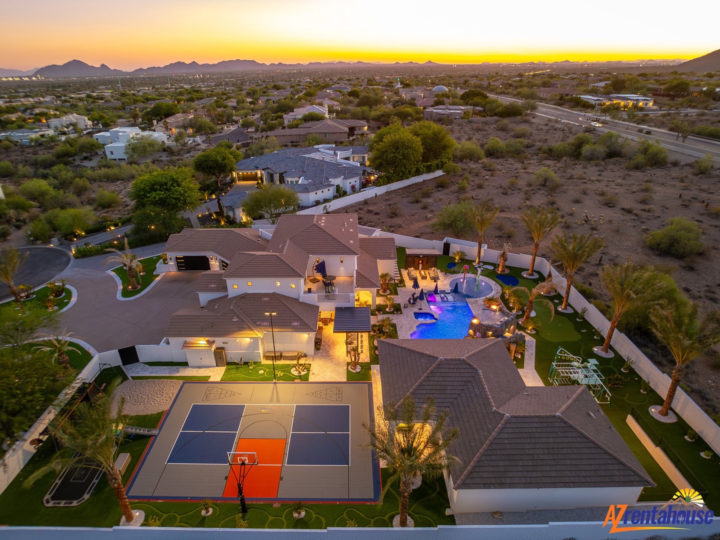 Stunning desert estate featuring expansive grounds, sparkling pool, basketball court, and panoramic mountain views at sunset.