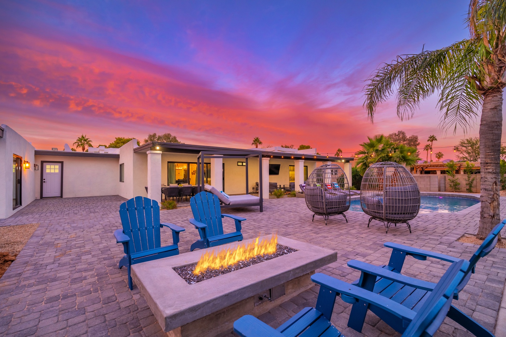 A stunning desert retreat framed by swaying palms and vibrant sunset skies, featuring a cozy fire pit, inviting egg chairs, and sparkling pool for warm evenings under the stars.