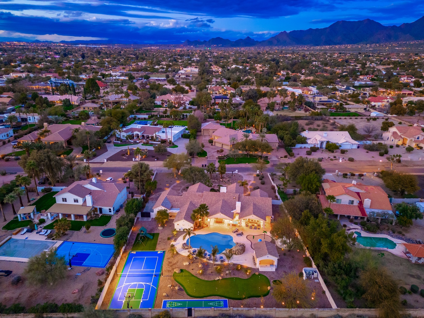 Aerial view of an expansive desert estate at twilight, featuring a swimming pool, sports courts, and manicured grounds surrounded by mountain vistas and neighboring properties.