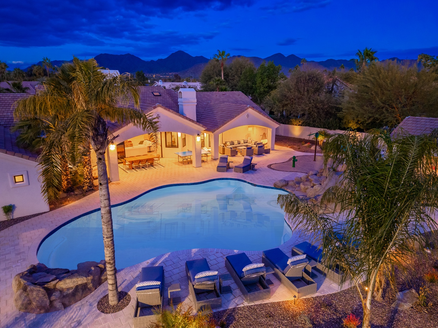 Aerial twilight view showcasing a private desert retreat with pool, palm-lined patio, and mountain backdrop—an inviting oasis in a peaceful residential neighborhood.