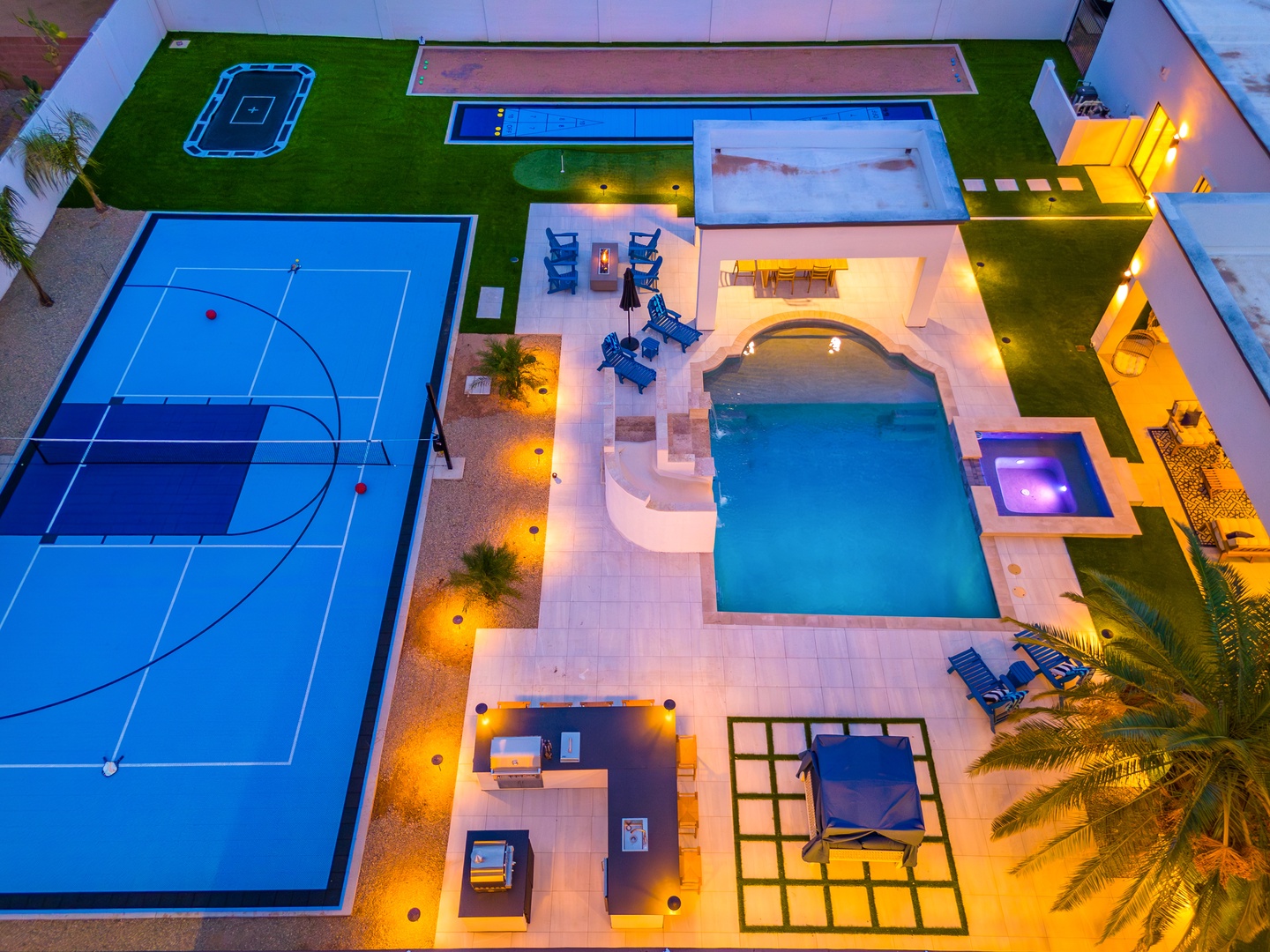 Aerial view showcasing the property's extensive outdoor amenities, including a full-size basketball court, swimming pool with curved slide, hot tub, and multiple lounging areas surrounded by lush green turf and tropical palms.