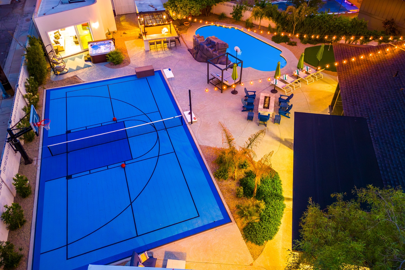 Aerial view of the property showcasing a private basketball court, outdoor swimming pool, covered patio, and beautifully landscaped grounds illuminated at night.