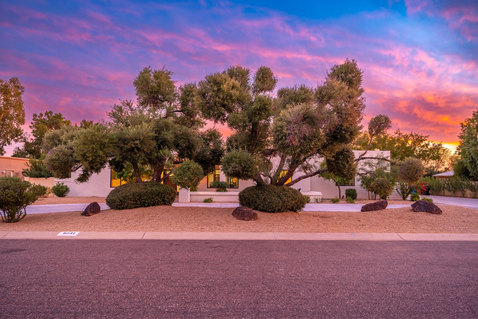 A striking desert property framed by mature trees and desert landscaping, set beneath a dramatic sunset sky painted in vibrant pink and orange hues.