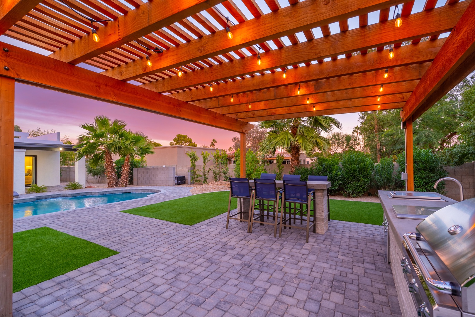 Your private backyard oasis features a stunning pergola with ambient lighting, perfect for outdoor dining. Enjoy the sparkling pool, manicured lawn, and BBQ area under a breathtaking desert sunset.