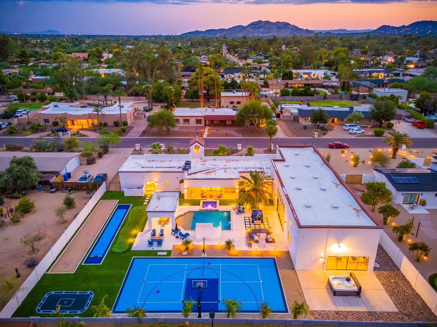 Aerial view of a resort-style property at dusk, featuring dual pools, sport courts, and expansive outdoor areas set against a peaceful neighborhood and mountain backdrop.