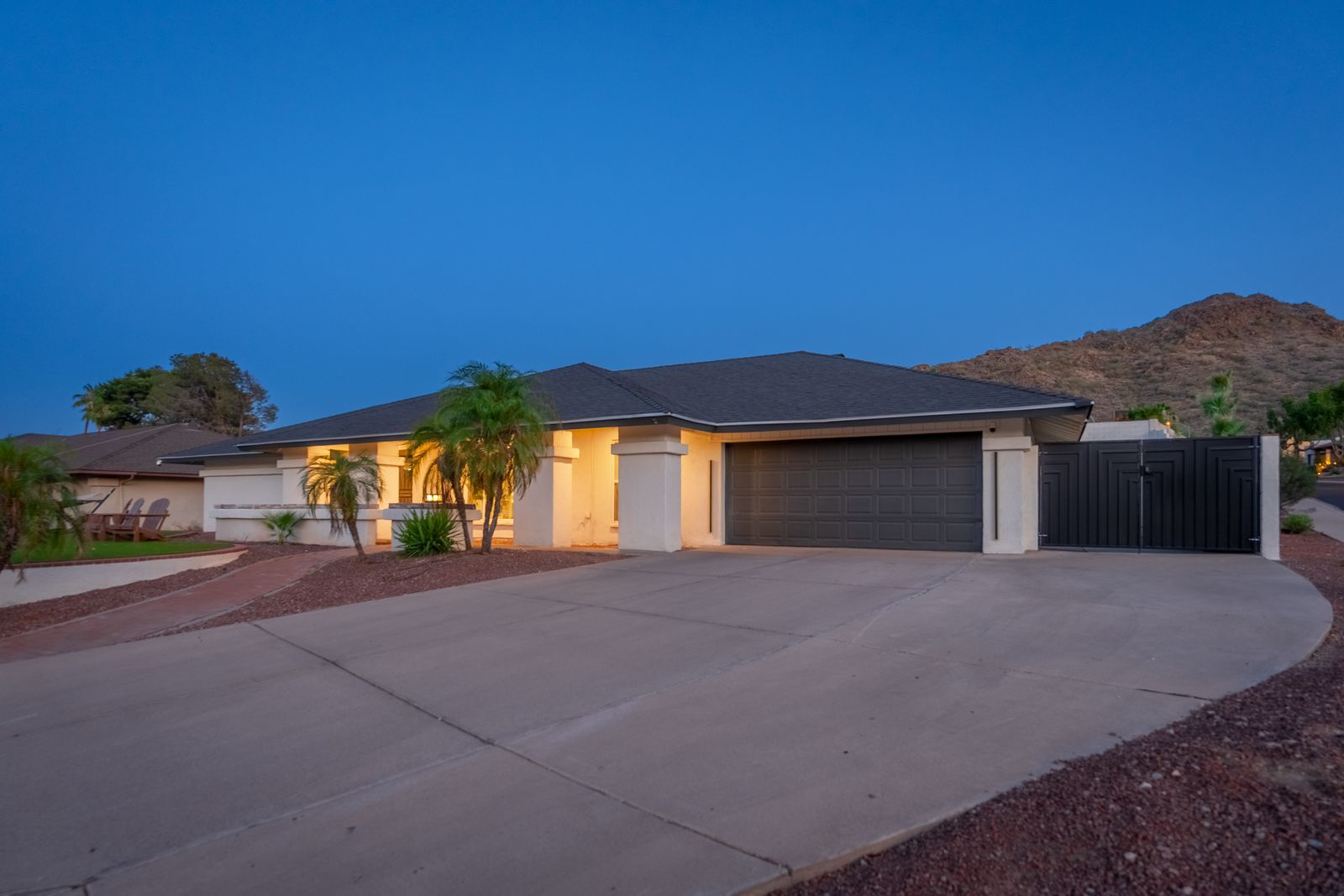 Desert oasis property with illuminated entryway, set against rugged mountain backdrop as twilight settles over the quiet neighborhood—a peaceful retreat awaits your arrival.