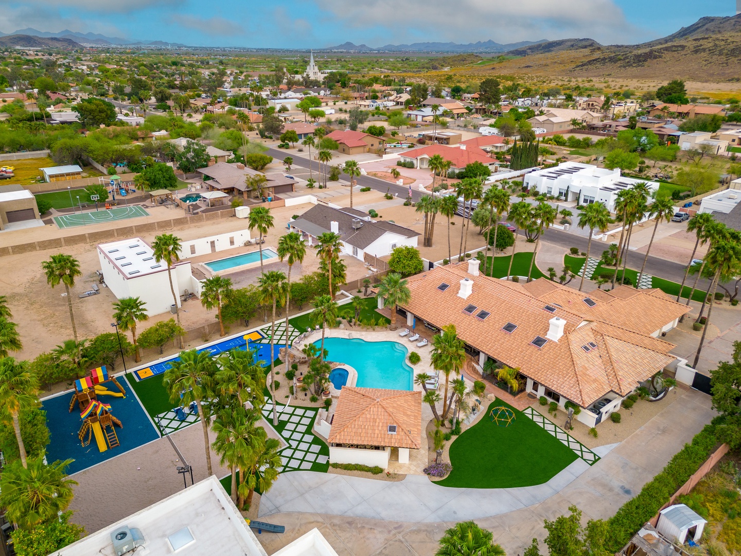 Aerial view of a vacation rental property featuring a sparkling pool, playground, tennis court, and lush landscaping set in a scenic mountain-framed neighborhood.