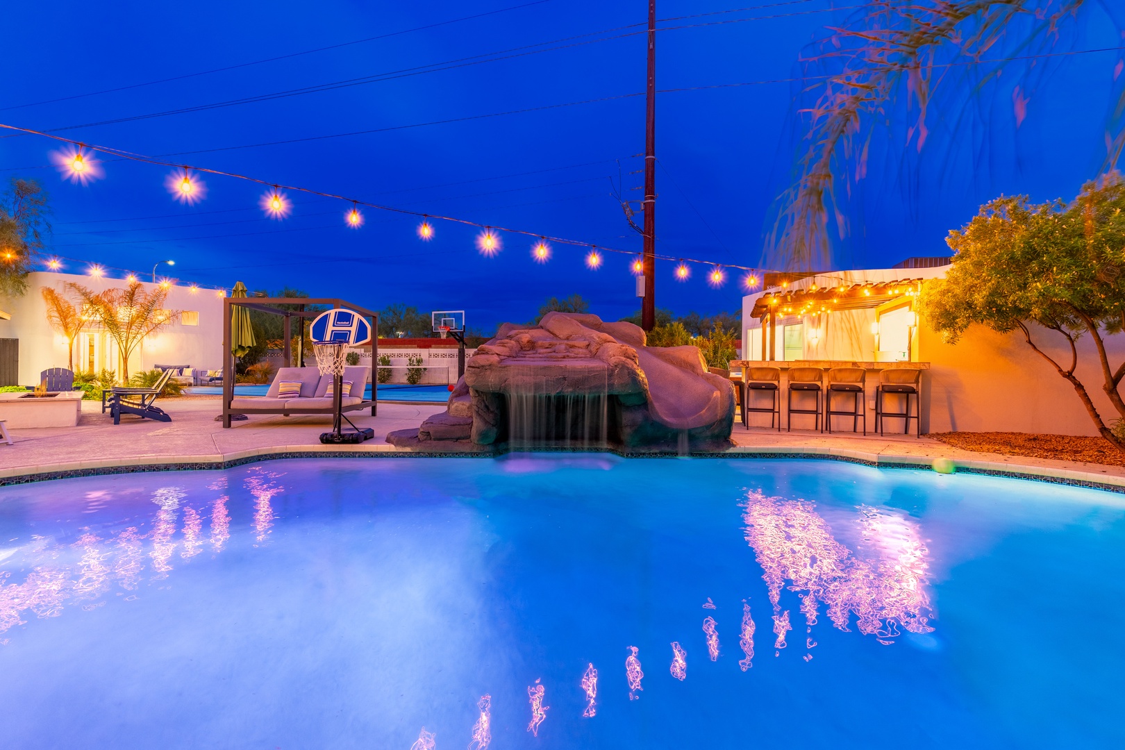 Twilight transforms the backyard into a glowing retreat. String lights reflect off the pool, while a rock waterfall and outdoor bar set the stage for unforgettable evenings under the stars.