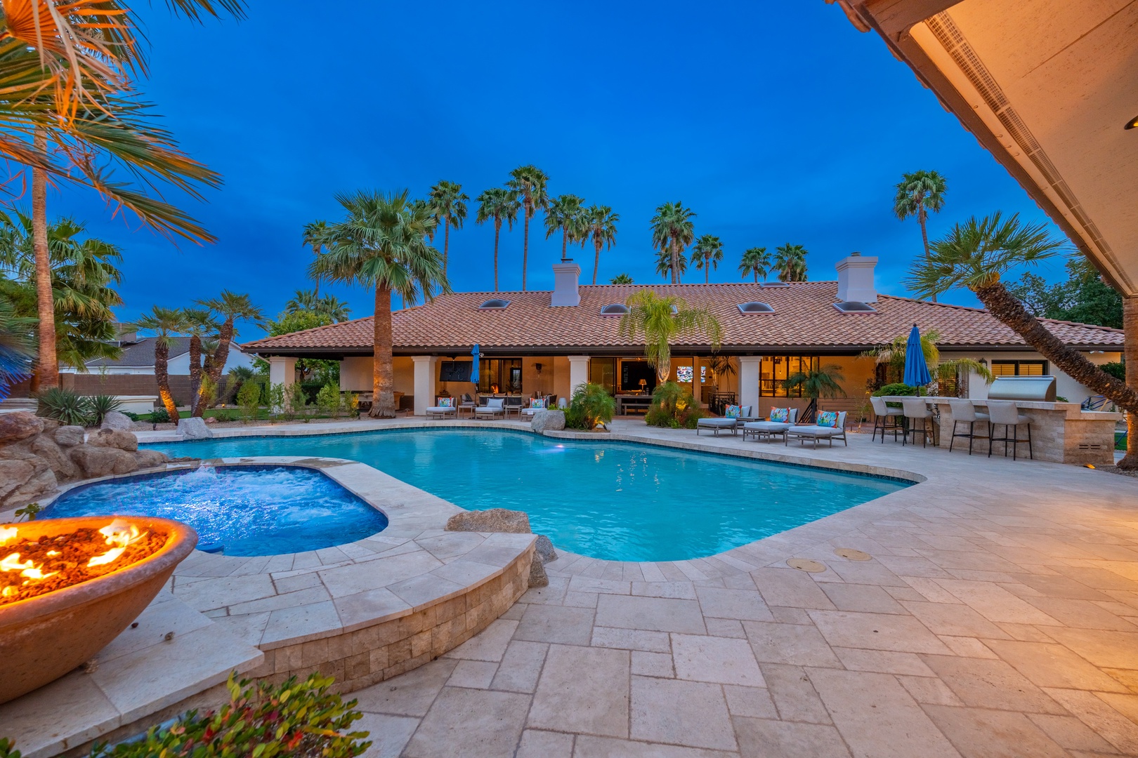 A sprawling backyard oasis features a stunning pool and spa surrounded by swaying palms, glowing fire features, and poolside lounging under the twilight sky.