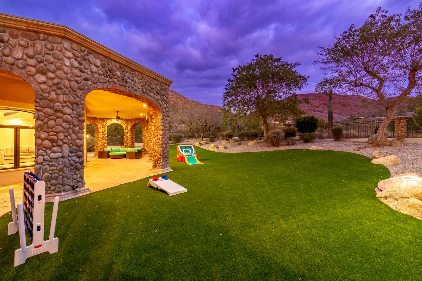Majestic desert villa at twilight, surrounded by natural landscape and mountains, featuring illuminated stone archways and a spacious lawn perfect for outdoor games and gatherings.