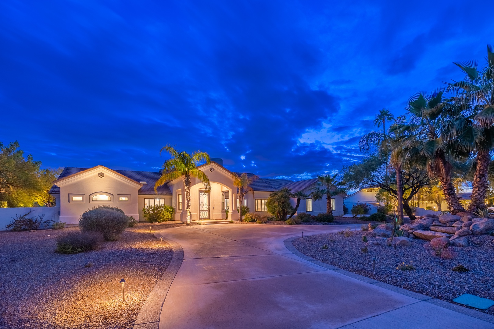 Elegant desert retreat framed by towering palms and native landscaping, captured at dusk with dramatic blue skies overhead.