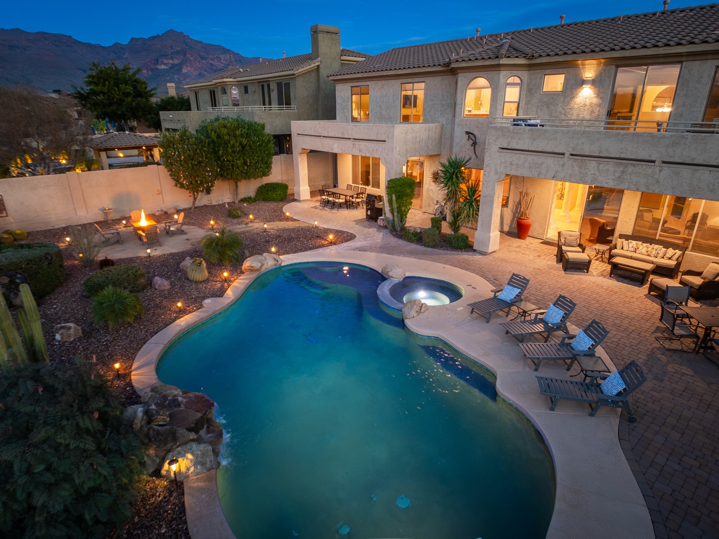 Twilight reveals a stunning two-story retreat with a sparkling pool, bubbling hot tub, and landscaped backyard nestled beneath dramatic mountain peaks—where desert evenings invite relaxation and unforgettable memories.