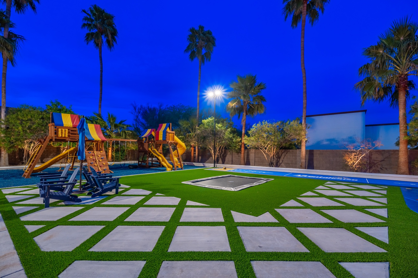 A resort-style backyard featuring a vibrant playground, lush artificial turf, and elegant palm trees beneath twilight skies. Perfect for family gatherings and outdoor fun.
