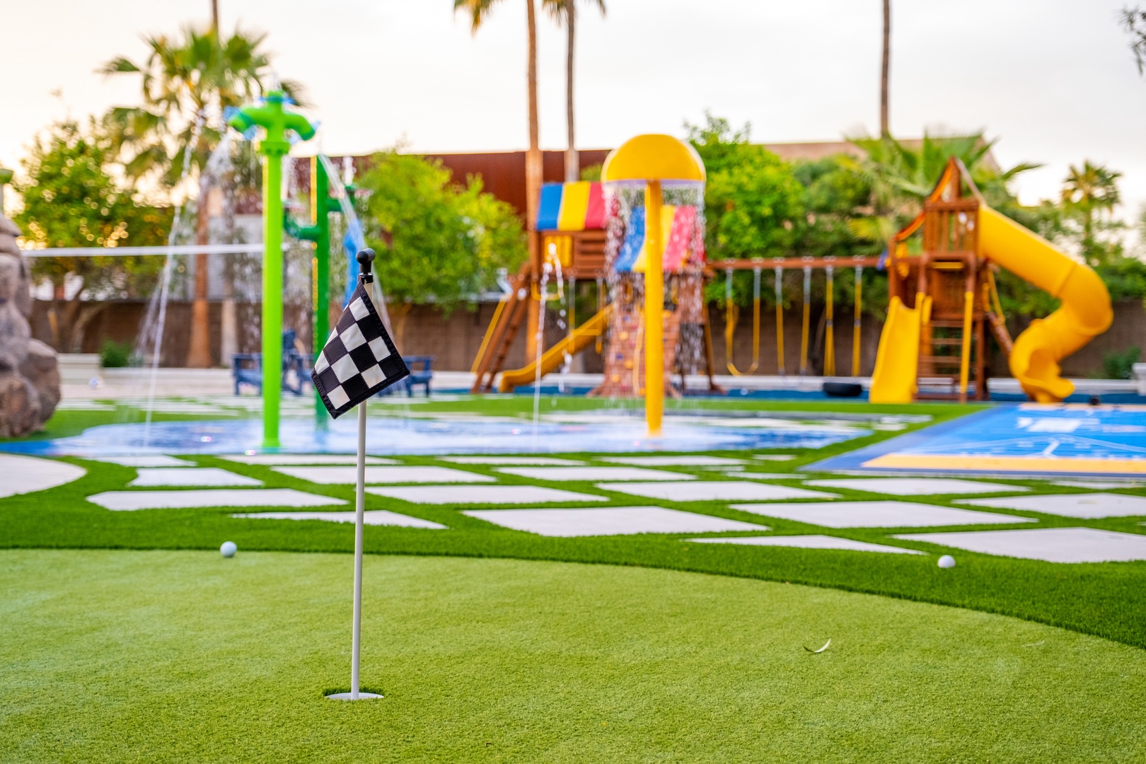 A family-friendly outdoor play area featuring a miniature putting green, vibrant water features, playground equipment, and slides surrounded by palm trees and lush greenery.