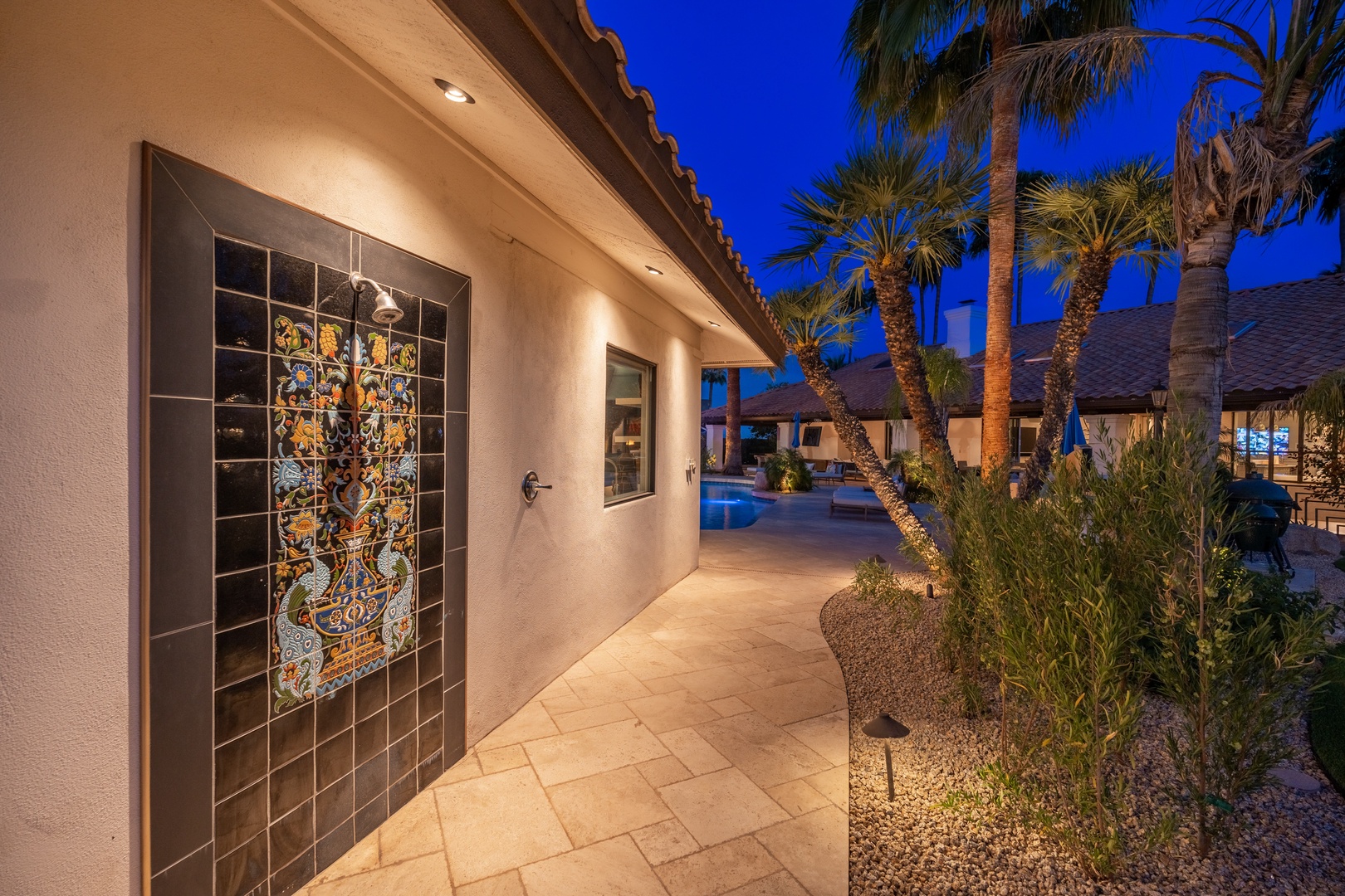 Warm lighting highlights the entrance featuring decorative tilework, with palm trees and pool area creating a resort-style backdrop at twilight.