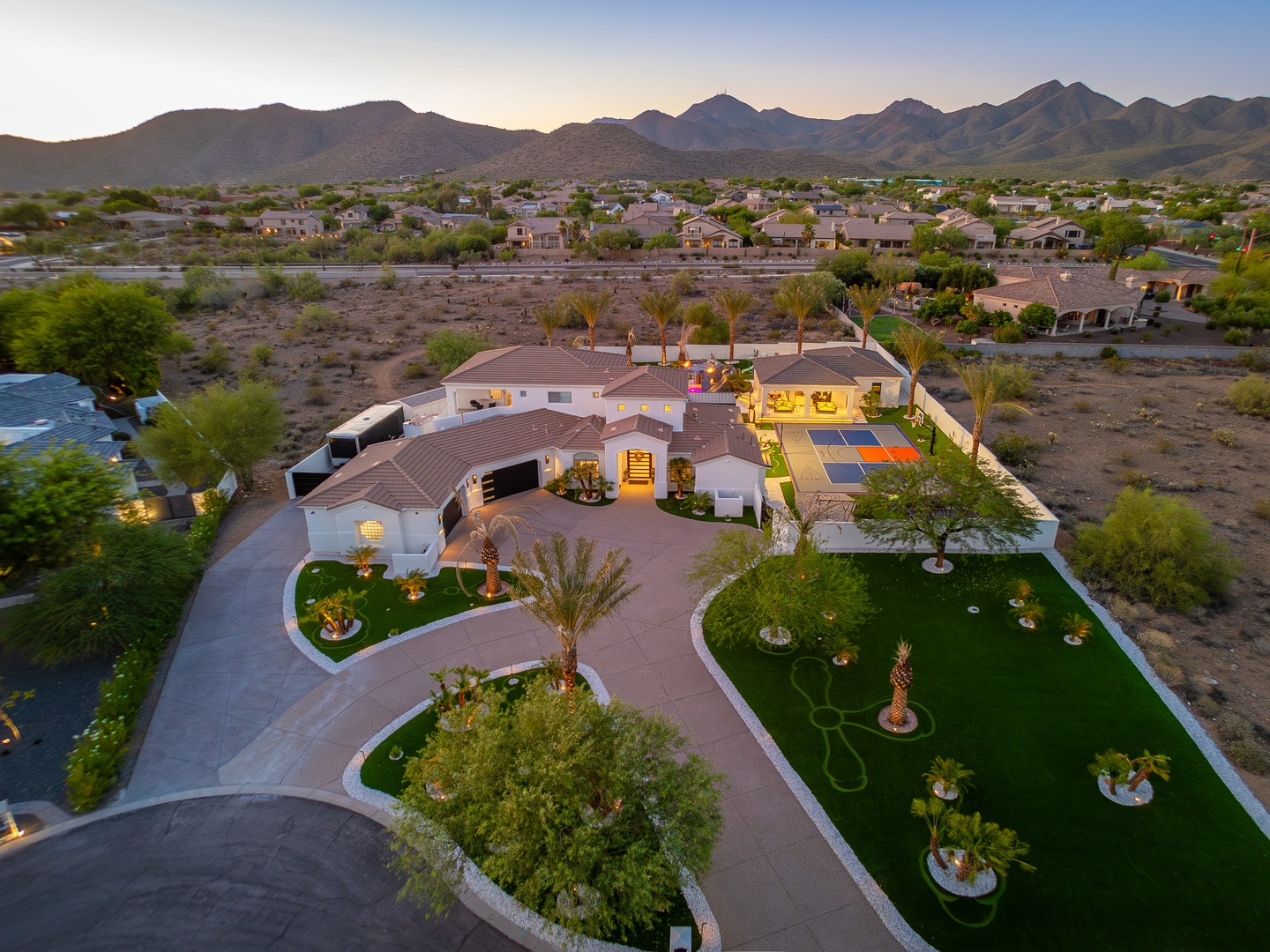 Luxurious desert estate nestled in a private neighborhood with dramatic mountain backdrop and expansive landscaped grounds featuring sport courts and palm-lined gardens at twilight.