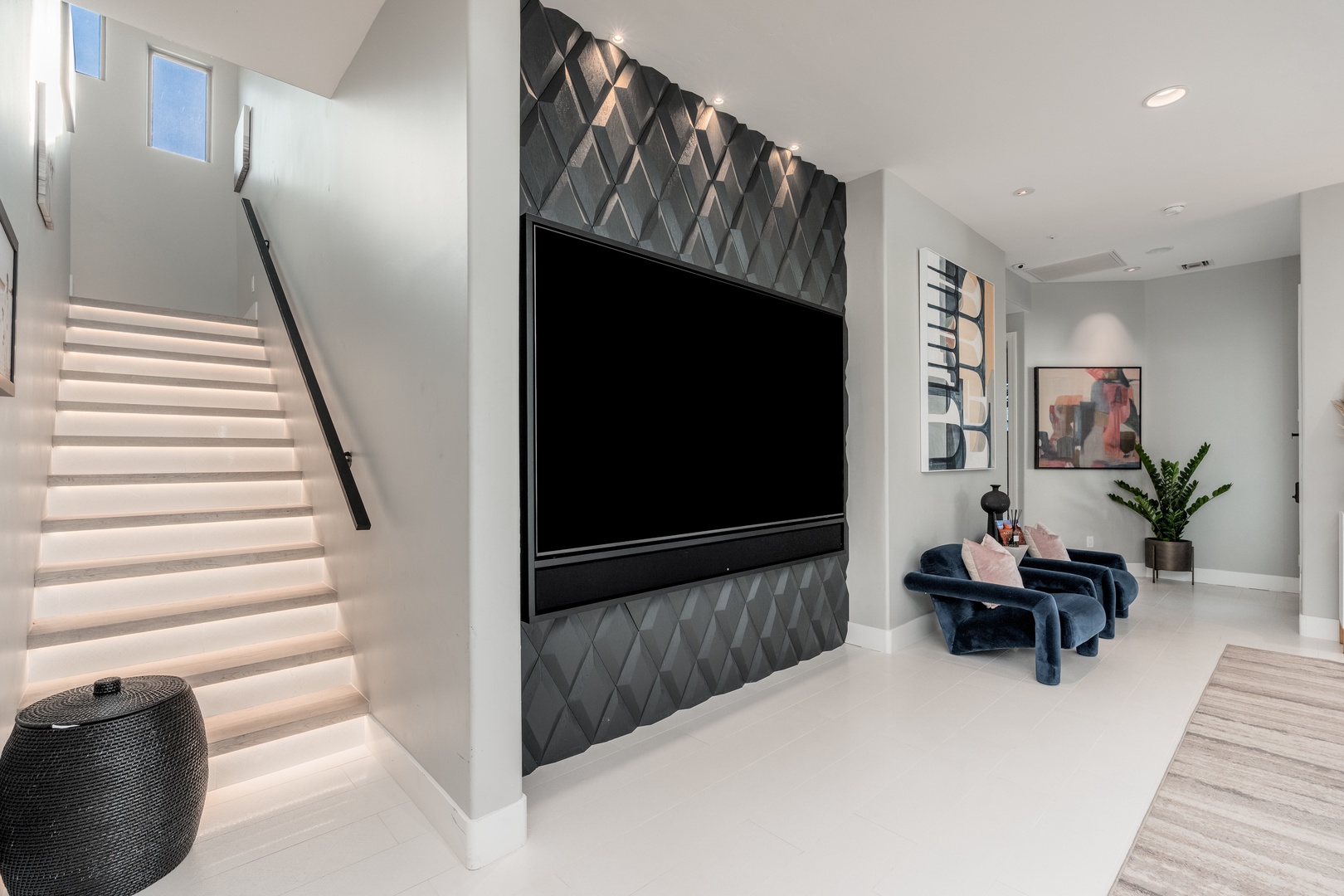 A striking living space features a textured accent wall framing your entertainment center, plush designer seating, and modern artwork. Illuminated stairs create a dramatic architectural element inviting you upstairs.