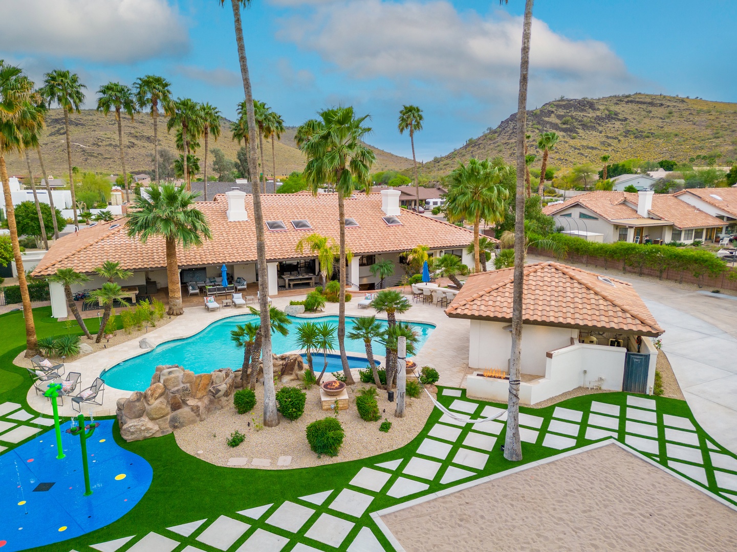Aerial view of a Spanish-style vacation home featuring a private resort-like backyard with a tropical pool, towering palms, children's play area, and mountain vistas in a sunny desert setting.
