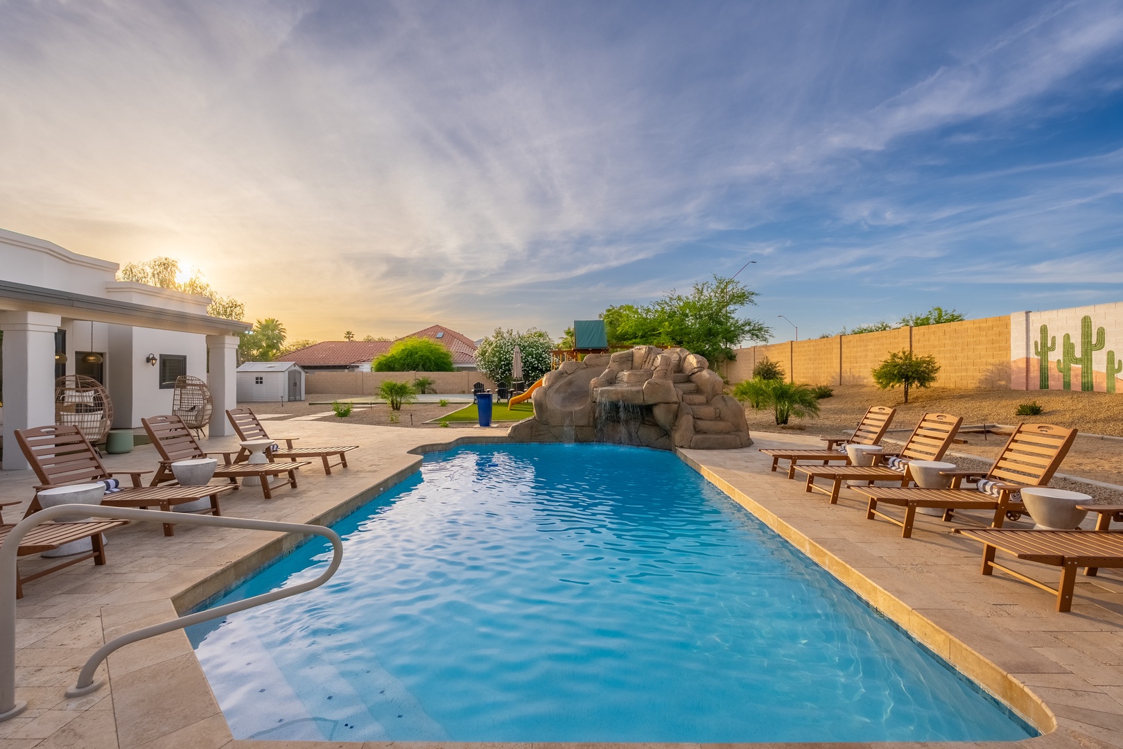 A spacious desert oasis awaits, with a sparkling pool, rock waterfall, and ample loungers framing a vibrant Arizona sunset.