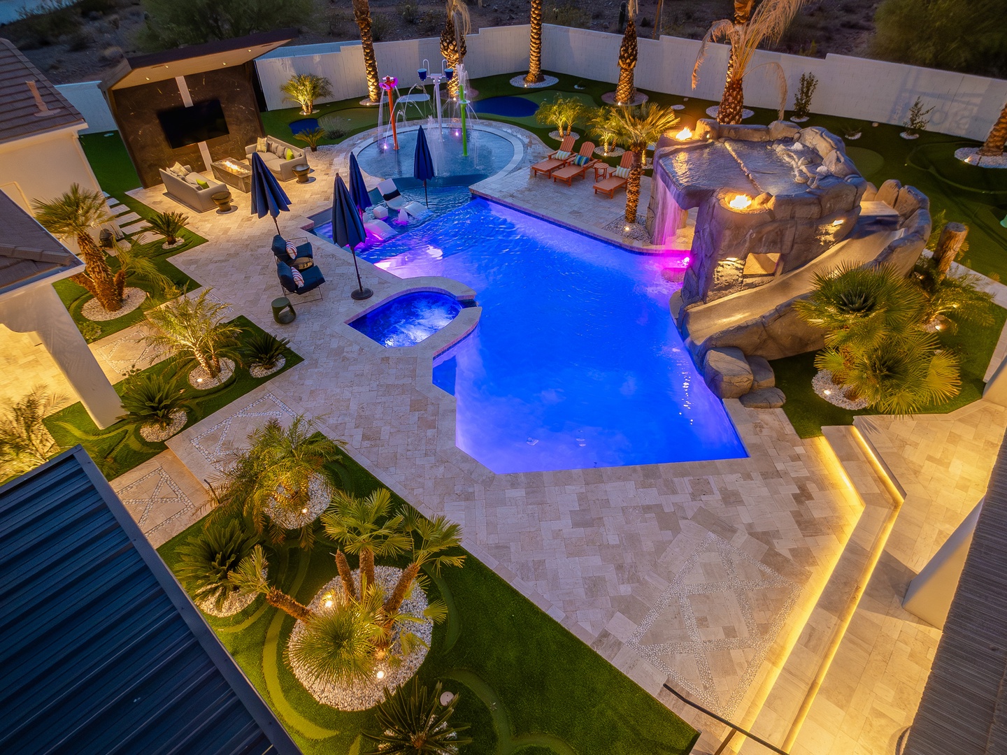 Aerial view of a stunning resort-style pool complex with cascading water features, illuminated palms, and rock formations set within a private walled compound at twilight.