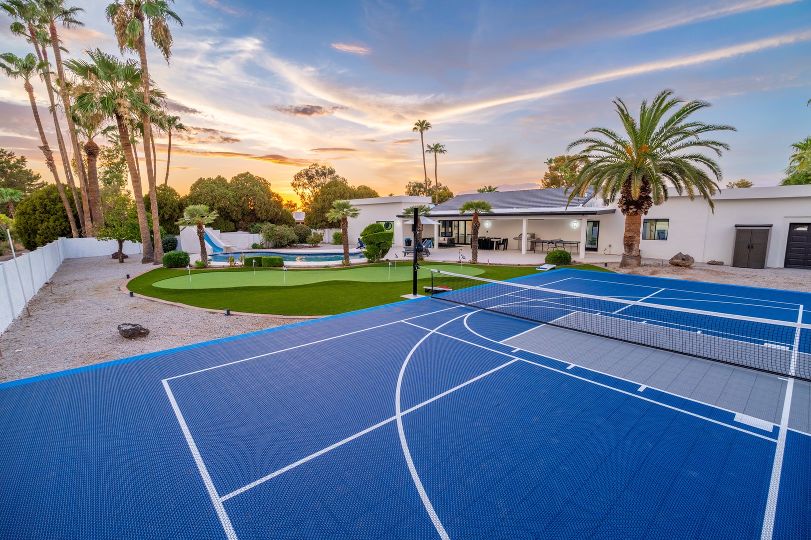 The property features a vibrant blue sport court, lush putting green, sparkling pool, and tropical palms—perfect for active days and relaxed evenings under stunning desert sunsets.