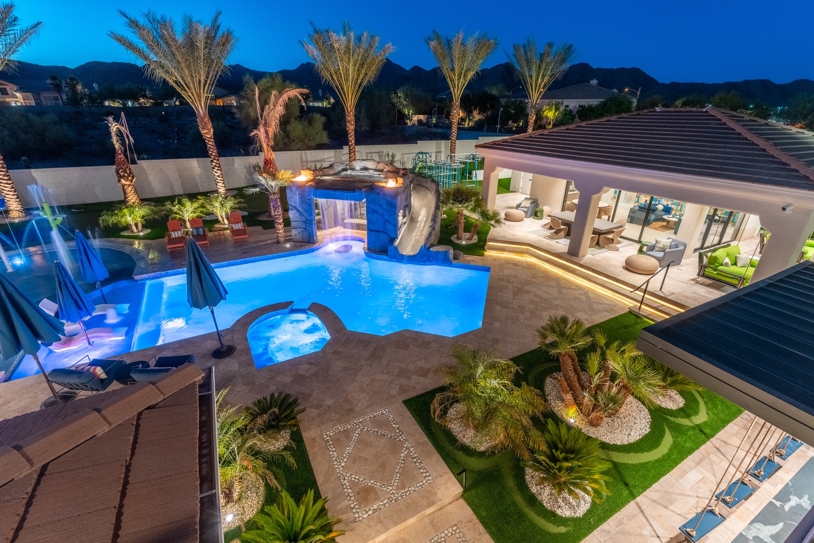 An aerial view of the property reveals a resort-style backyard oasis with illuminated pool featuring a waterslide and grotto, palm tree landscaping, covered patio, and mountain backdrop at twilight.