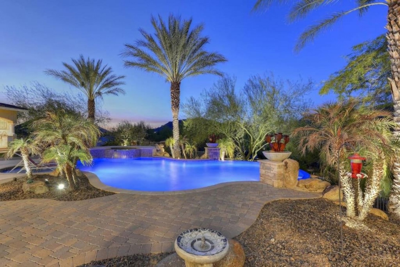 A stunning desert resort-style pool glowing blue at twilight, surrounded by palm trees, decorative landscaping, and mountain views—an elegant outdoor oasis in a peaceful natural setting.
