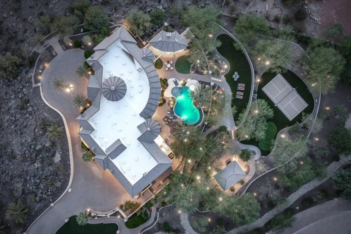 Aerial view reveals the expansive property nestled in desert landscape, featuring a glowing pool, tennis court, and extensive grounds surrounded by natural terrain.