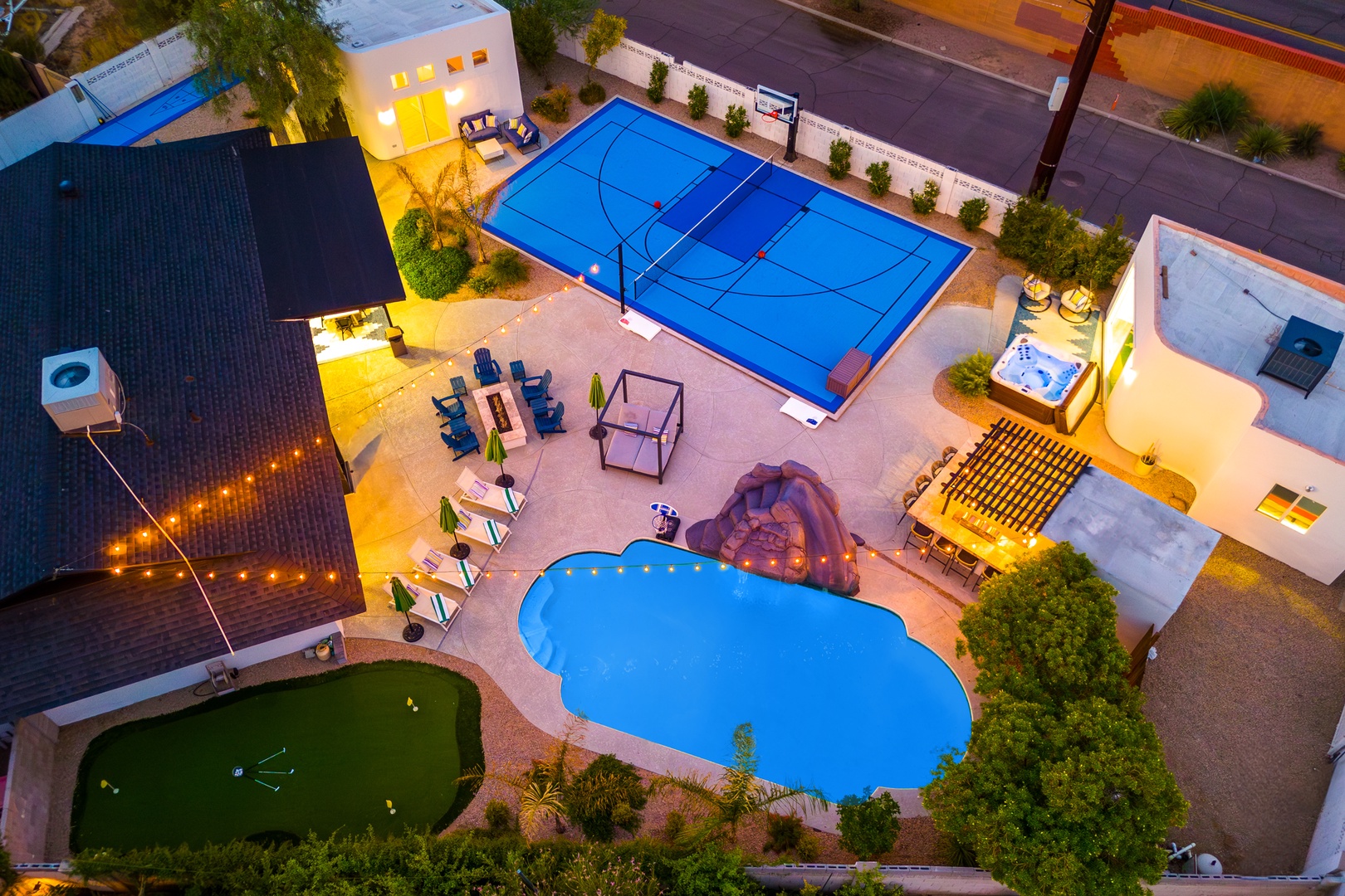 Aerial view showcasing an upscale vacation rental property featuring a freeform swimming pool, sport court, putting green, outdoor dining areas, and multiple covered lounging spaces, set in a residential neighborhood at twilight.