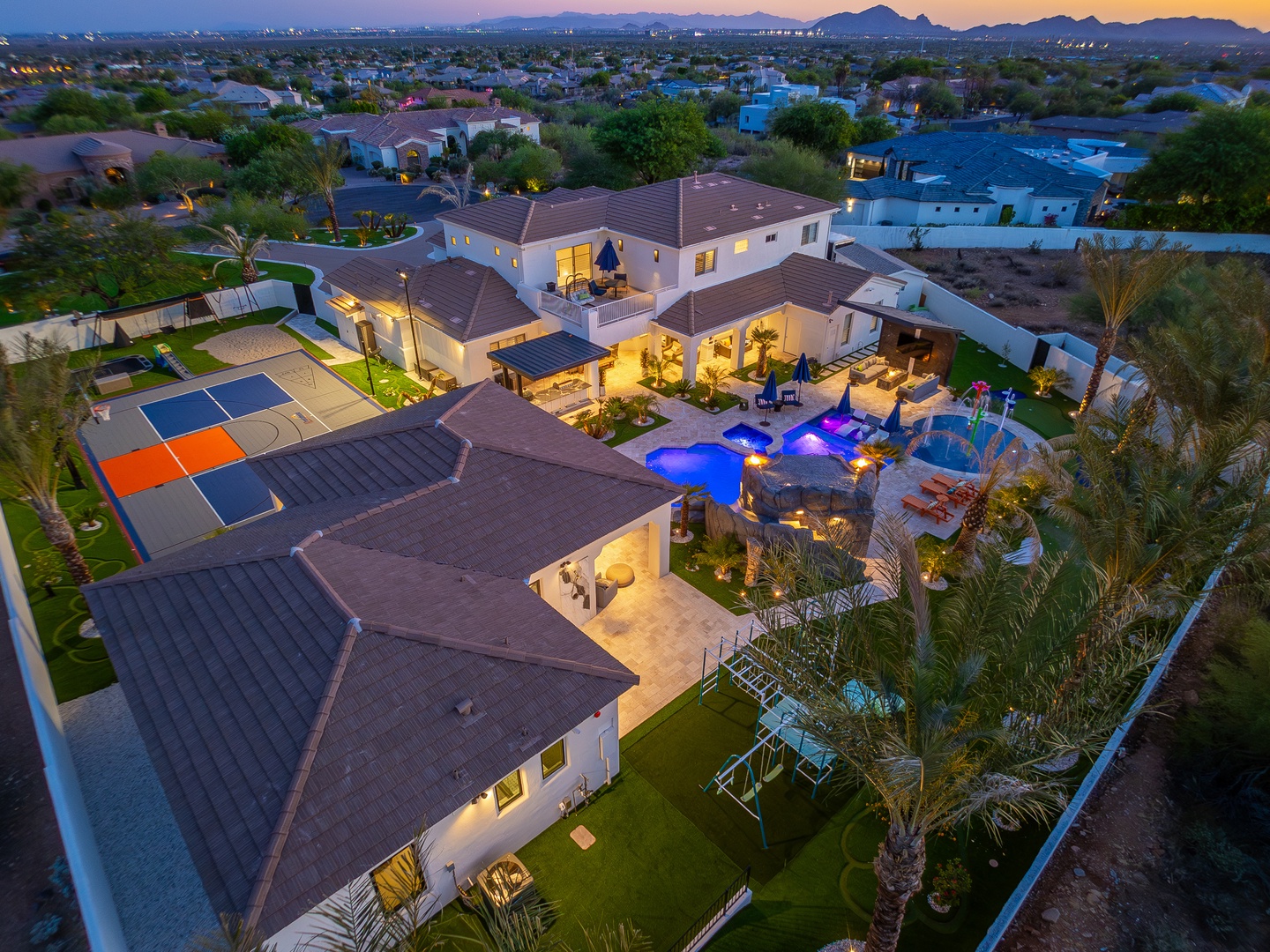 Aerial view of a sprawling desert estate featuring a resort-style pool, sports court, and lush landscaping, set against a mountain backdrop at twilight.