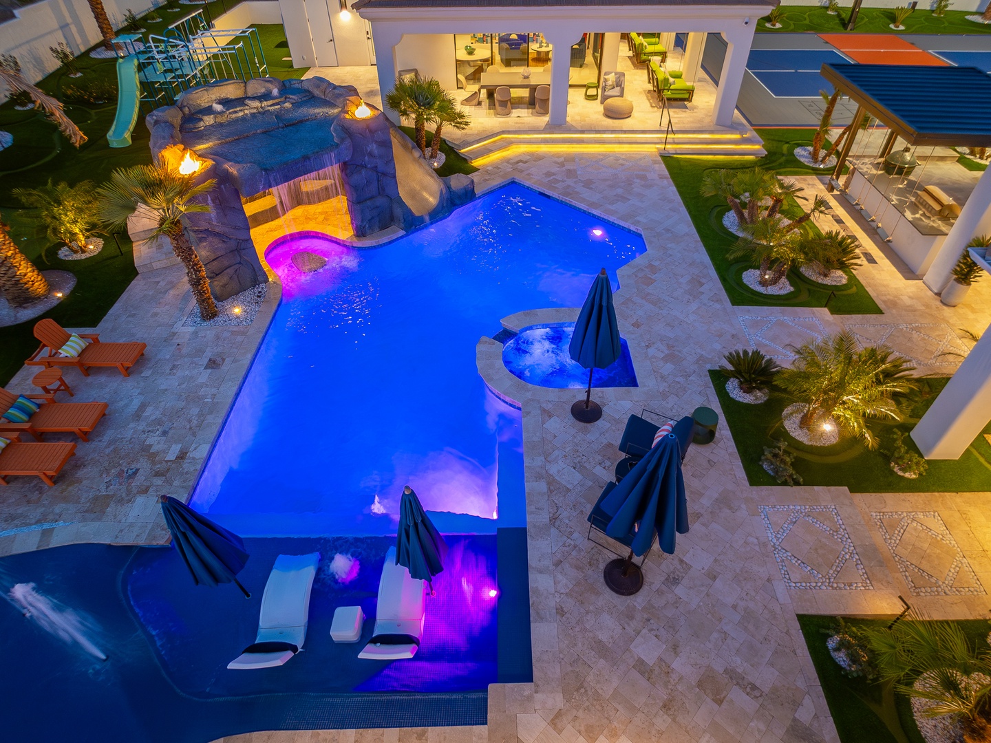 Aerial view of luxury property featuring illuminated pool with colorful lighting, hot tub, outdoor lounge areas, and modern architectural design at dusk.
