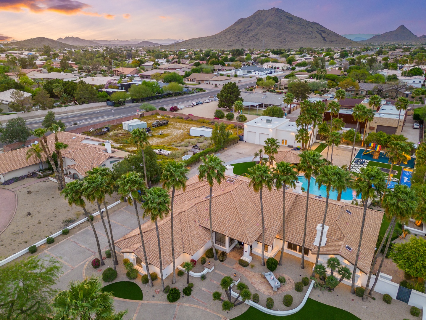 Aerial view of the property nestled in a desert neighborhood, framed by dramatic mountain peaks at sunset. Lush palm trees line the entrance to a spacious estate with a private pool and play area.